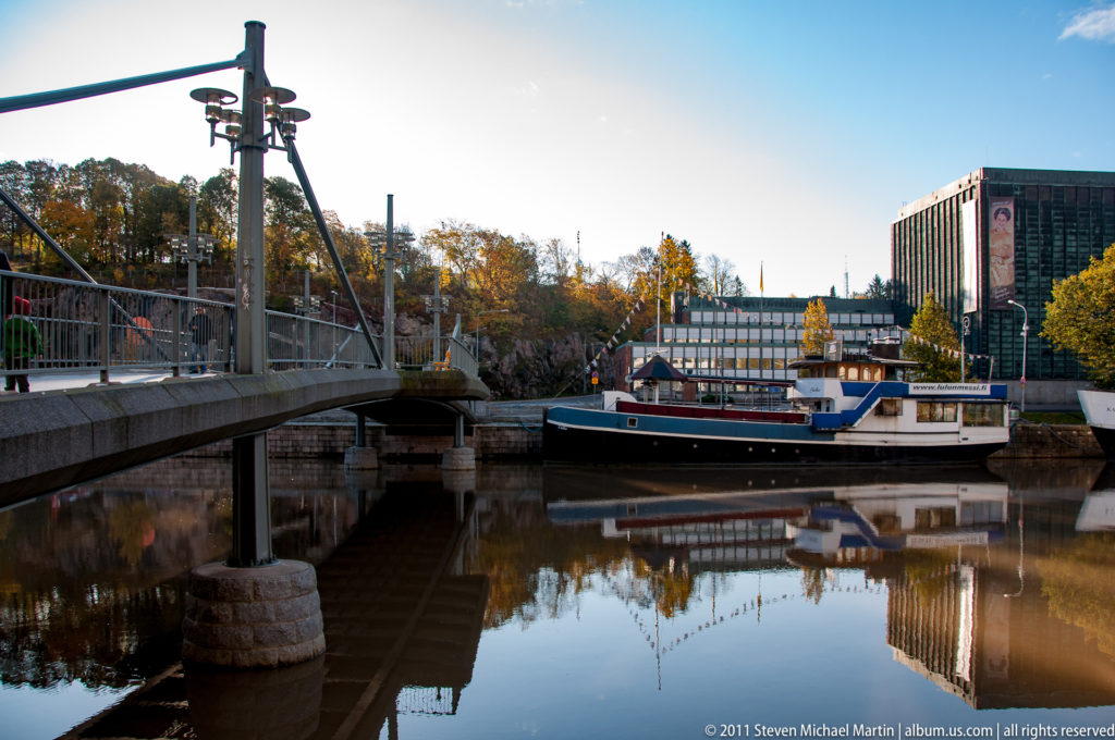 Low pedestrian bridge crossing a calm canal beside a moored blue-and-white boat, with buildings reflected in the water.