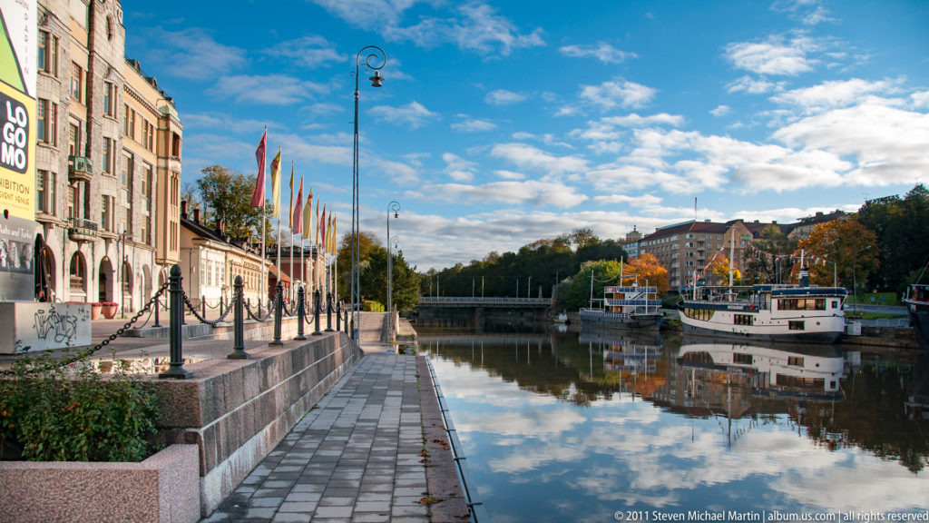 Stone canal promenade with flags and lamps beside calm water, moored boats, a bridge, and buildings reflected under a blue sky.
