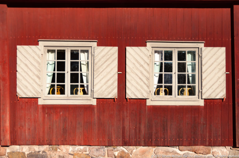 Two white multipane windows with diagonal-board shutters set into a weathered red wooden wall above a stone foundation.