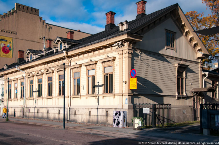 Pale wooden corner house with tall ornamented windows, bracketed eaves, chimneys, and a dark roof in Old Rauma.