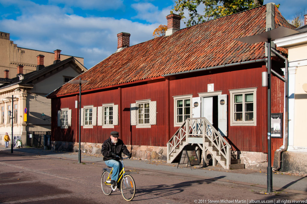 Cyclist riding past a long red wooden house with white windows, tiled roof, stone base, and exterior entrance steps.