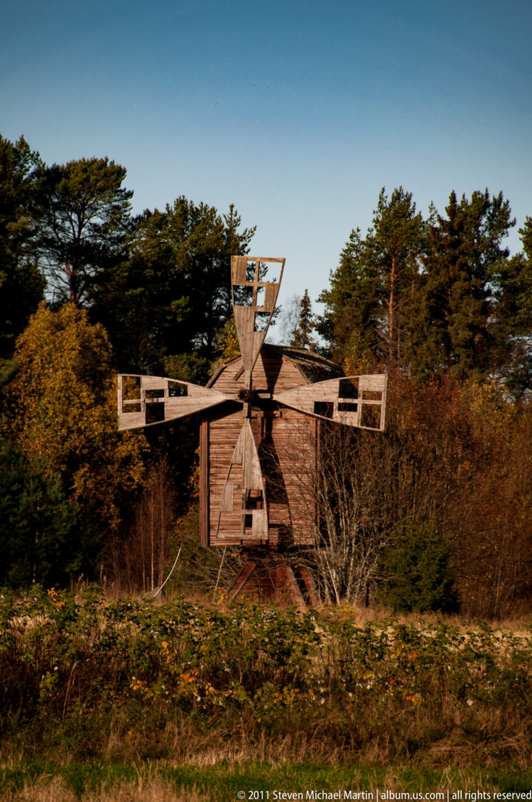 Weathered wooden windmill with four rectangular open-framed sails standing against dense trees in autumn light.