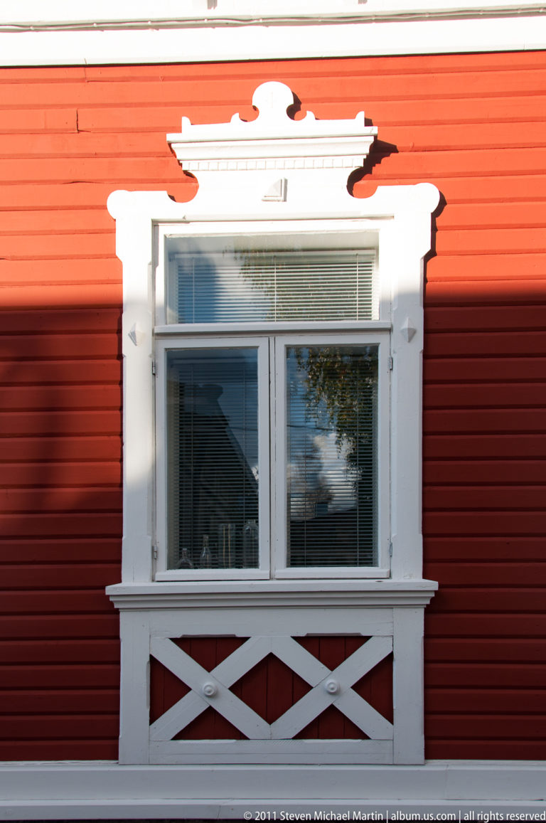 White ornamental window surround with shaped trim and cross-braced panel on a red horizontal-board wooden wall.