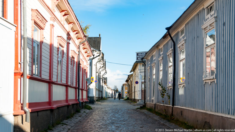 Narrow cobblestone street between red-and-white and gray wooden buildings beneath a clear blue sky in Rauma.