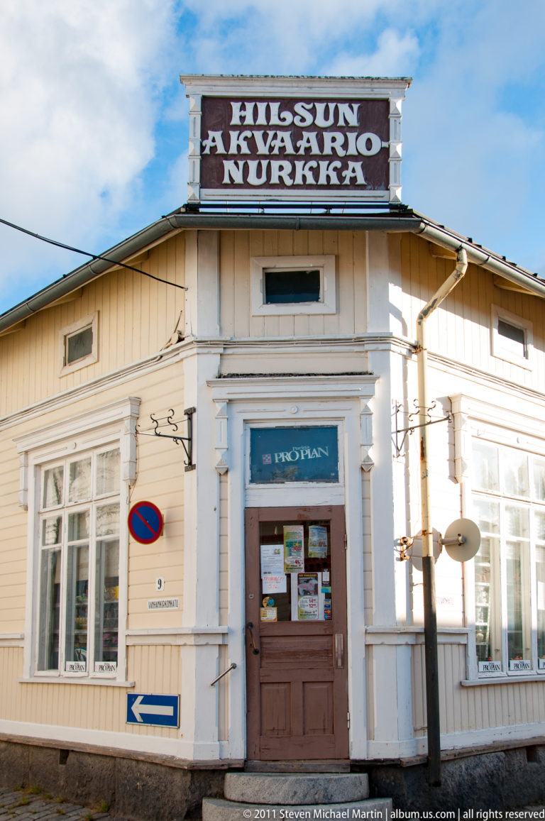 White wooden corner café building with large 'Huissun Aamukahvio Nurkka' rooftop sign, central brown door, and broad multipane windows.