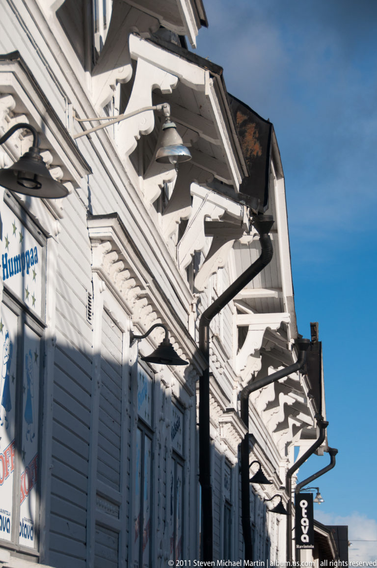 Upward view along a white wooden shopfront with repeated carved eave brackets, projecting signs, and layered trim against blue sky.