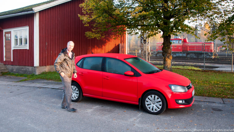Person stands beside a red hatchback near a red wooden building, with a locomotive and railway tracks behind.