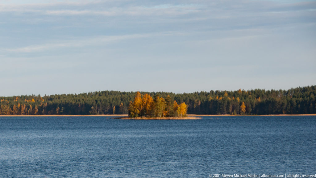 Broad blue lake with a small autumn-colored wooded island and dark forest stretching across the distant shore.
