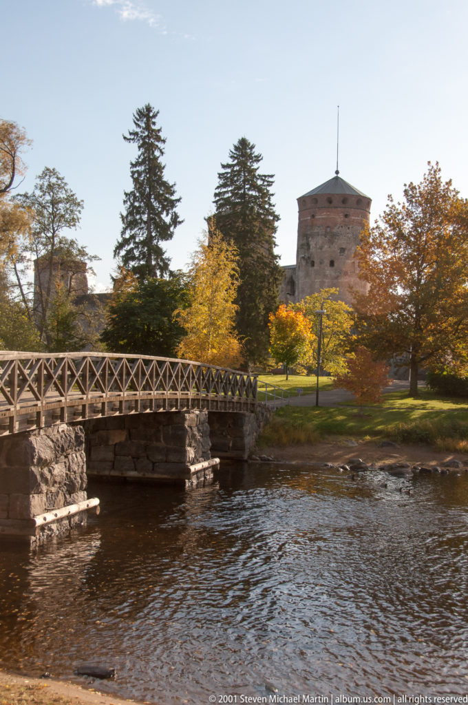 Wooden footbridge crosses a narrow channel toward autumn trees, with a round stone castle tower rising behind them.