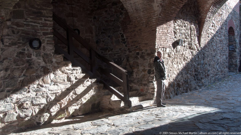 Rough-stone castle passage with a wooden stair at left and a visitor standing in a shaft of sunlight.