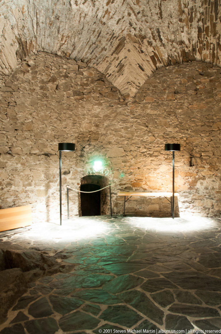Low stone-vaulted castle chamber with dark paved floor, spot lighting, barriers, and a small recessed opening.
