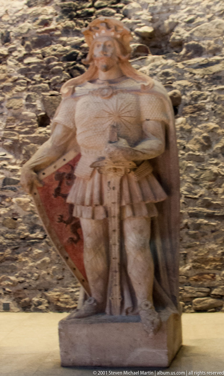 Blurred carved seated figure in historical dress displayed against a rough stone wall inside Olavinlinna Castle.