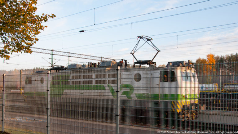 White-and-green electric locomotive behind a security fence with its pantograph raised to overhead catenary wires.