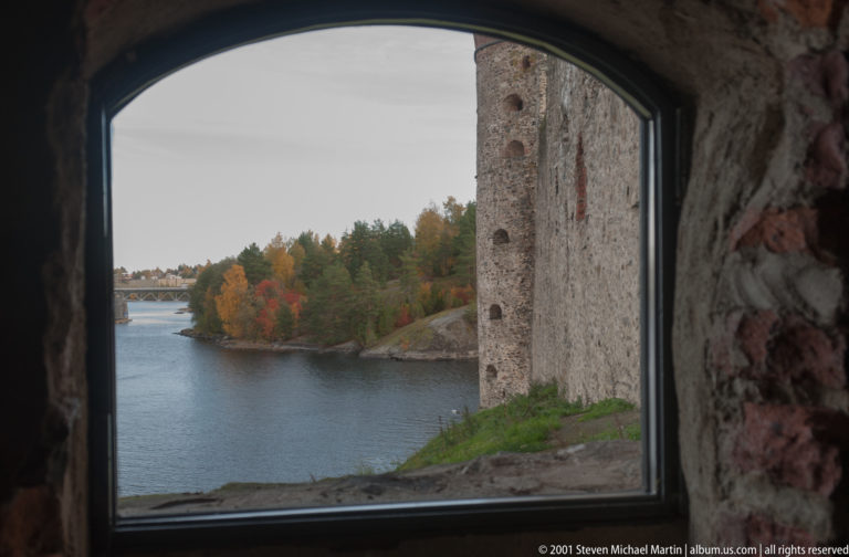 Autumn shoreline and water channel seen through a castle window, with a tall exterior masonry wall at right.