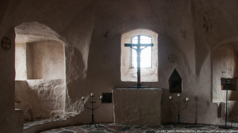 Small whitewashed castle chapel with crucifix before a narrow window, altar, candleholders, and stone floor.