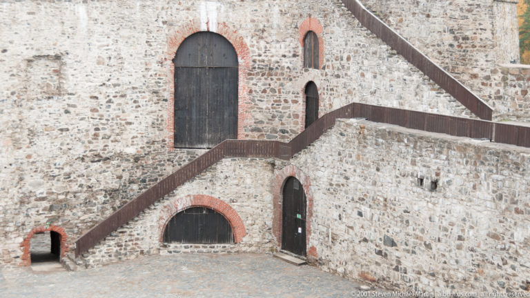 Zigzagging dark timber stairs crossing a stone castle wall with several red-brick-trimmed arched doorways.