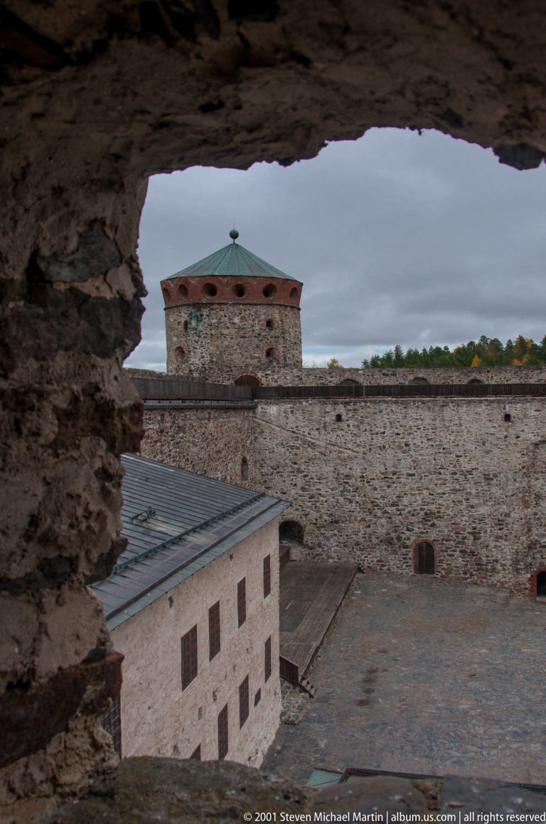 Inner castle courtyard, pale building, high stone wall, and round tower seen through a rough masonry opening.