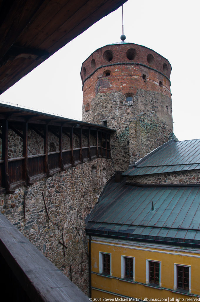 Round stone-and-brick castle tower rising above a timber gallery and ochre building with metal roof.