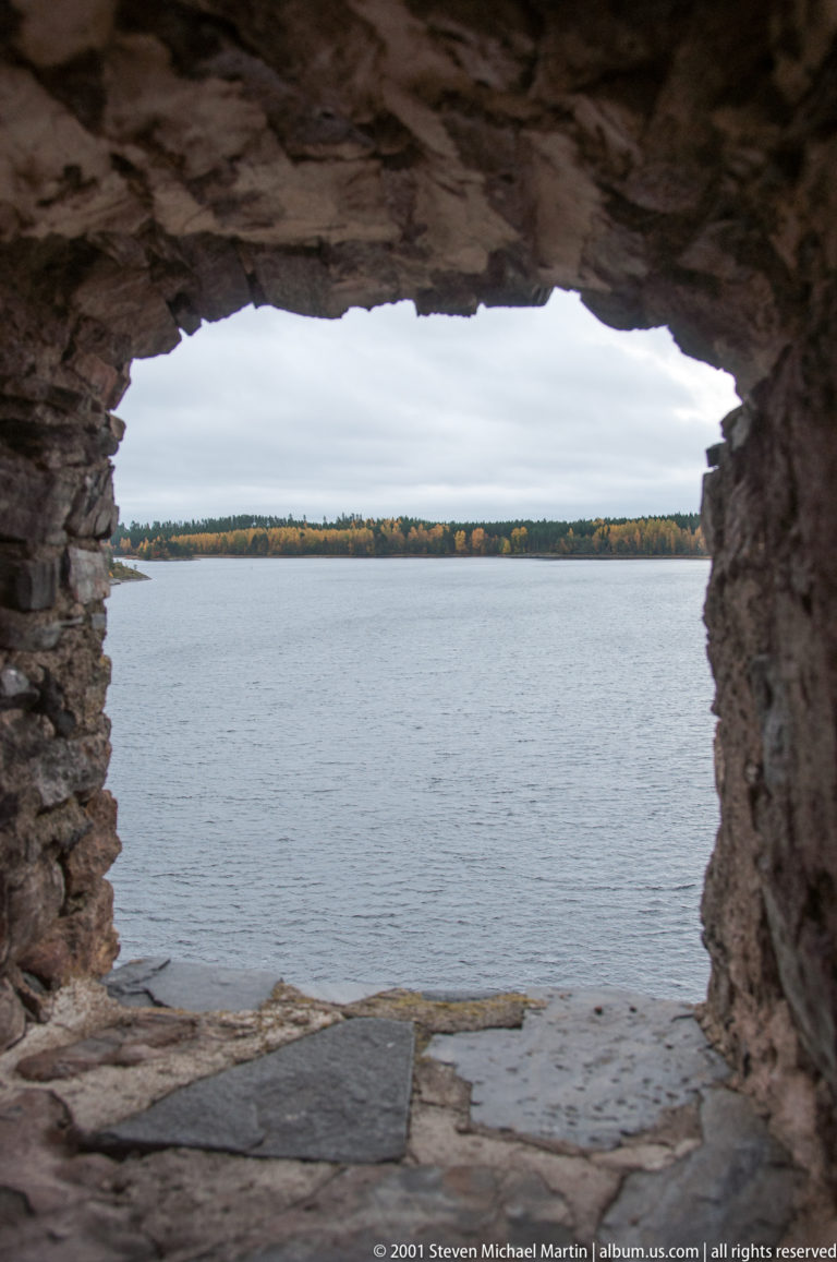 Gray lake and distant wooded shore framed by a broad rough stone opening in Olavinlinna Castle.