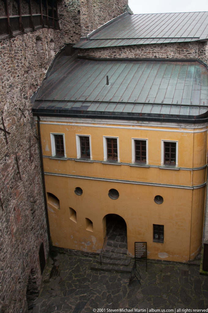 Ochre-painted castle building with small windows, arched doorway, and metal roof in a narrow stone courtyard.
