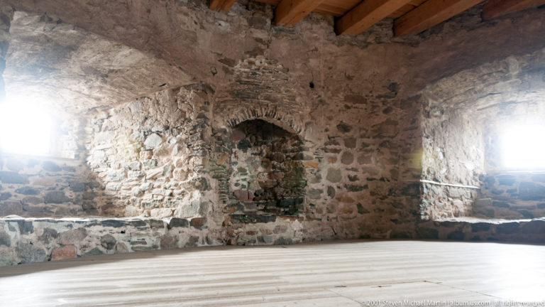 Empty stone castle chamber with deep windows, rough masonry, timber ceiling beams, and pale wooden floor.