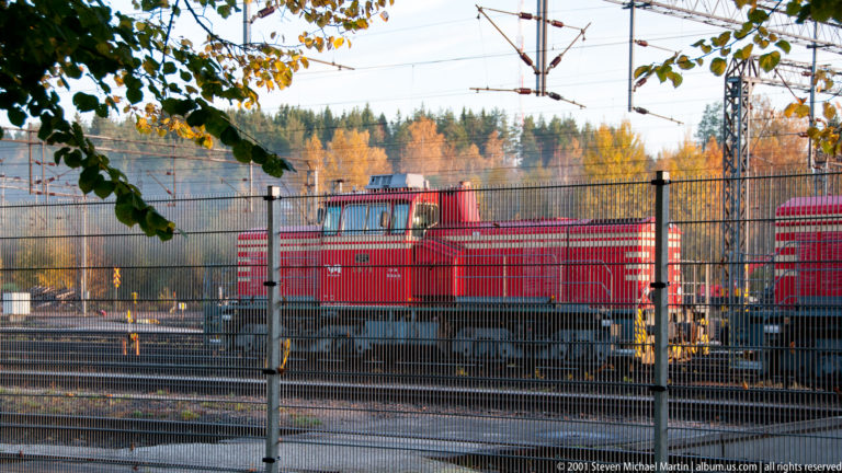 Red diesel locomotive behind a metal fence in a rail yard with overhead wires, tracks, and autumn trees.