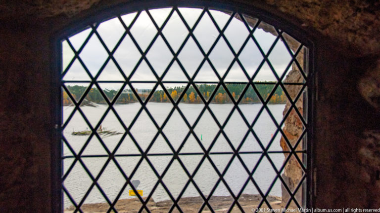 Lake and autumn shoreline viewed through a diamond-pattern iron grille in an arched castle window.