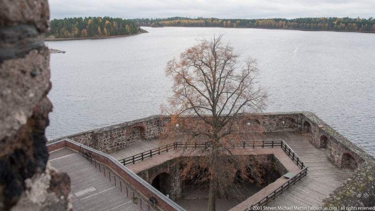 Leafless tree inside a walled castle terrace with timber railings and lake beyond.