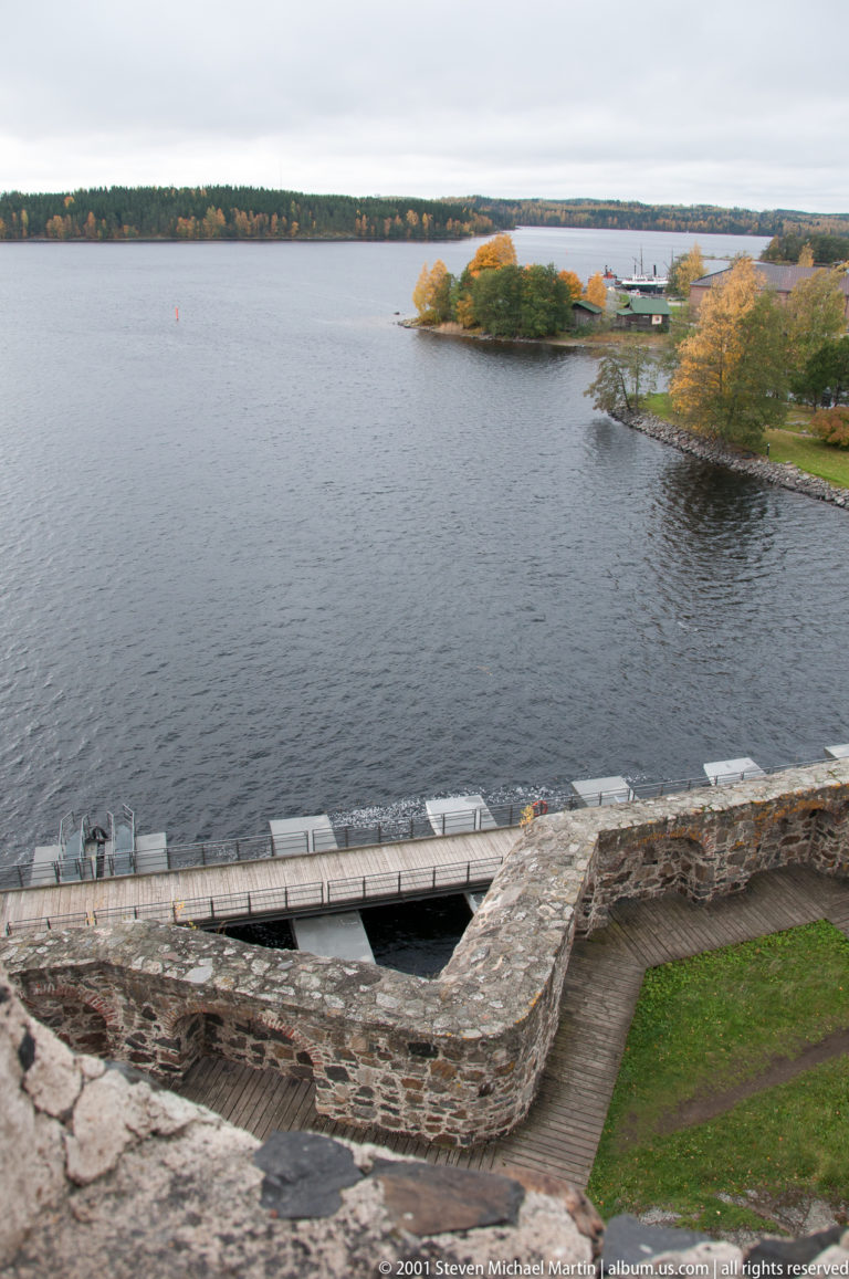 Elevated view over castle masonry and an access structure toward a wooded island across the lake.