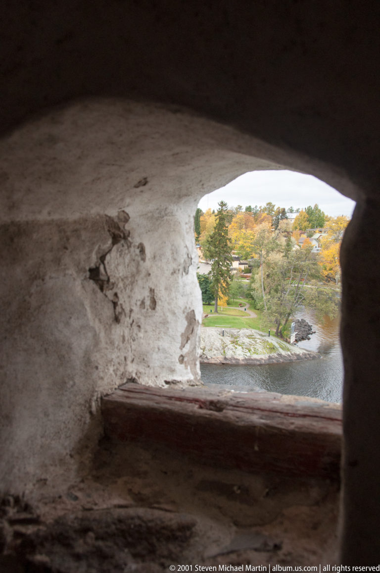 Autumn shoreline seen through a deep, rough-plastered masonry opening in Olavinlinna Castle.