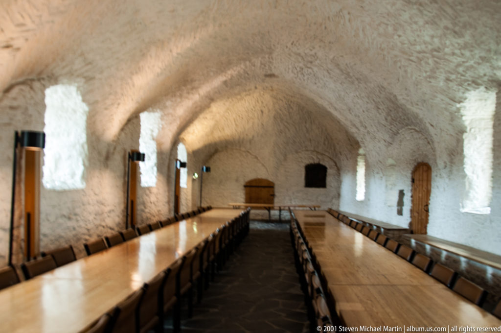 Long whitewashed vaulted castle hall with recessed windows, timber doors, and rows of tables and chairs.