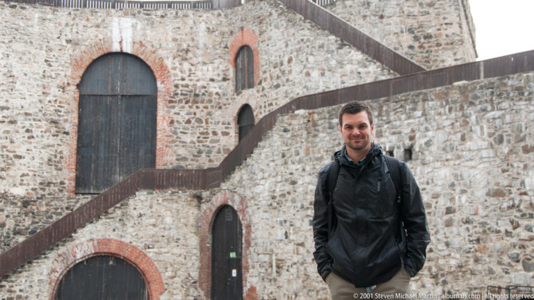 Man in dark jacket before rough stone masonry, brick-edged arches, and crossing timber stairs at Olavinlinna Castle.
