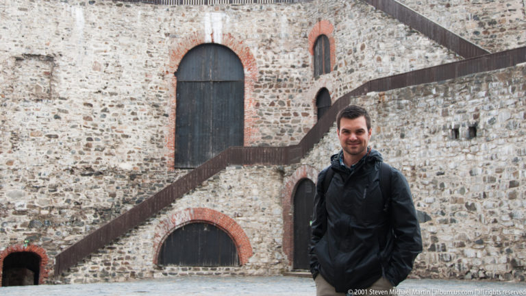 Man standing before a stone castle wall with brick-trimmed arches and diagonal dark timber stairways.