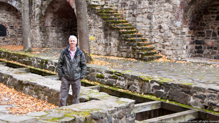 Visitor standing among low stone walls and a narrow channel beneath steep moss-covered castle steps.