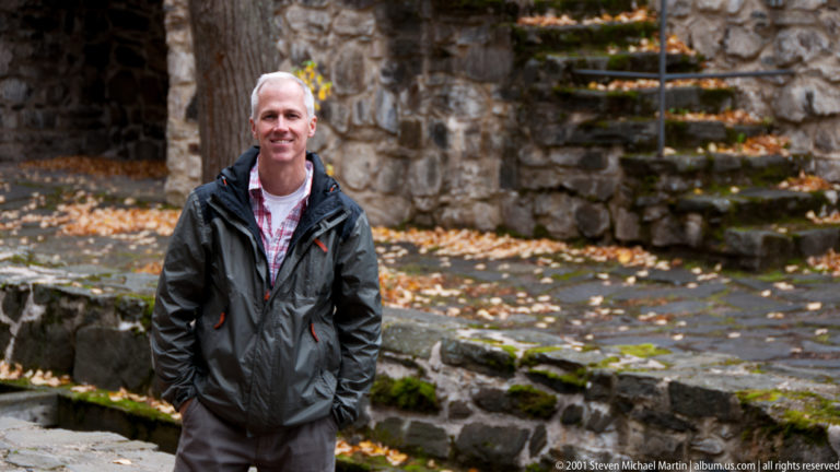 Man in dark jacket beside mossy stone steps, rough castle walls, paving, and fallen autumn leaves.