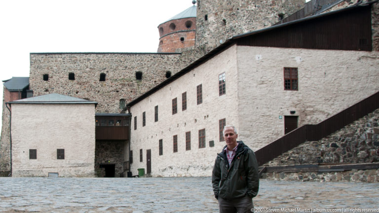 Man standing in a cobbled Olavinlinna Castle courtyard among whitewashed walls, stone masonry, stairs, and towers.