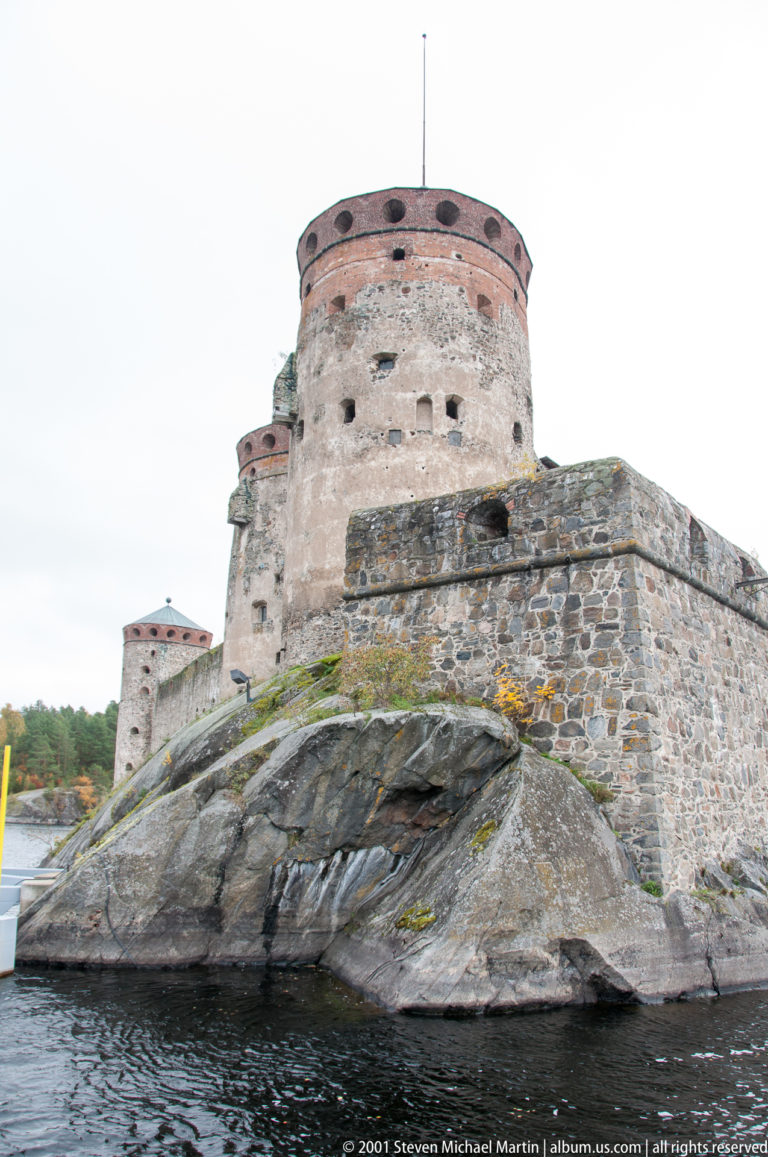 Round stone tower and curtain walls of Olavinlinna Castle rising from exposed bedrock beside dark lake water.
