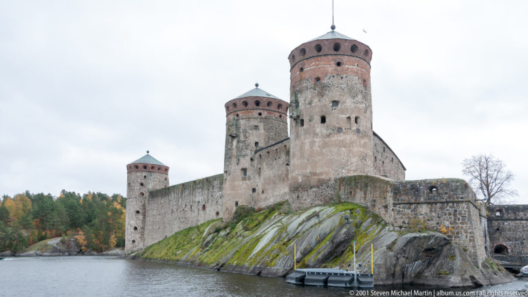 Three round towers and stone curtain walls of Olavinlinna Castle above exposed lakeside bedrock.