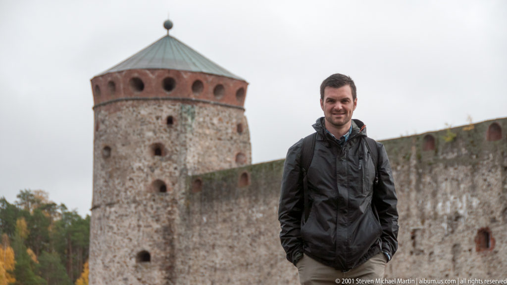 Man in dark jacket standing before a round stone-and-brick tower and curtain wall at Olavinlinna Castle.
