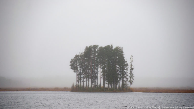 Small tree-covered island emerging from fog over a gray lake near Savonlinna.
