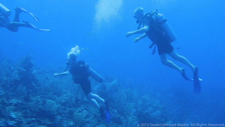 Two scuba divers swim at different depths above a coral reef while bubbles rise through blue water.