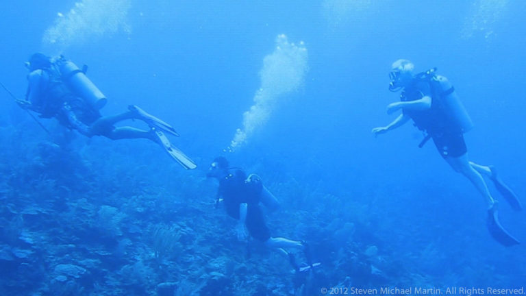Three scuba divers at different depths above a coral reef, with streams of bubbles rising overhead.
