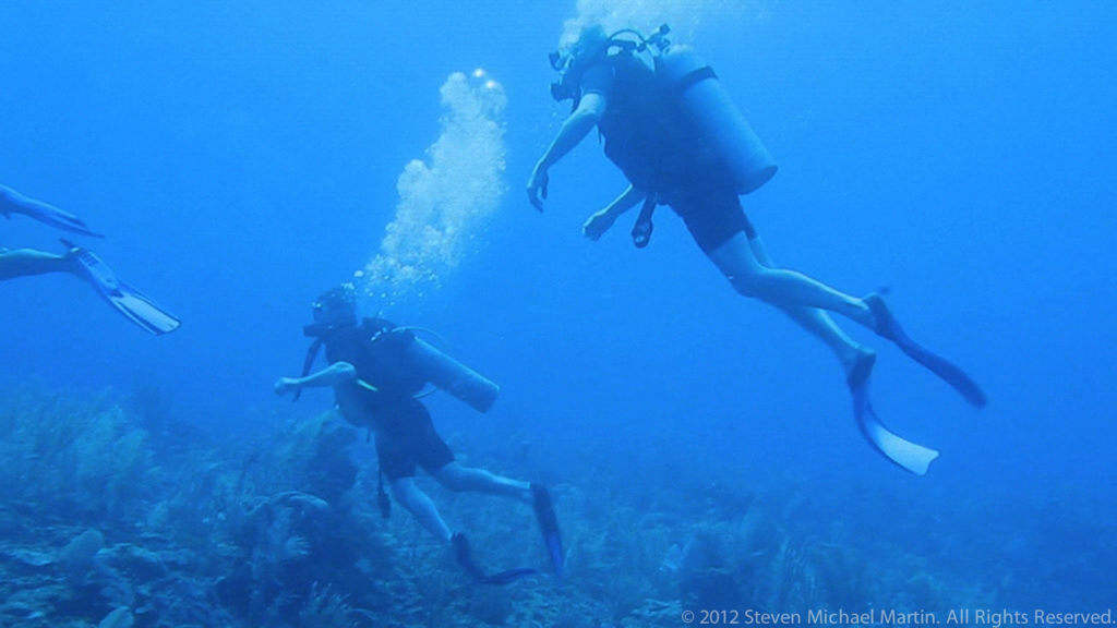 Two scuba divers angle through blue water above the reef, each trailing bubbles.