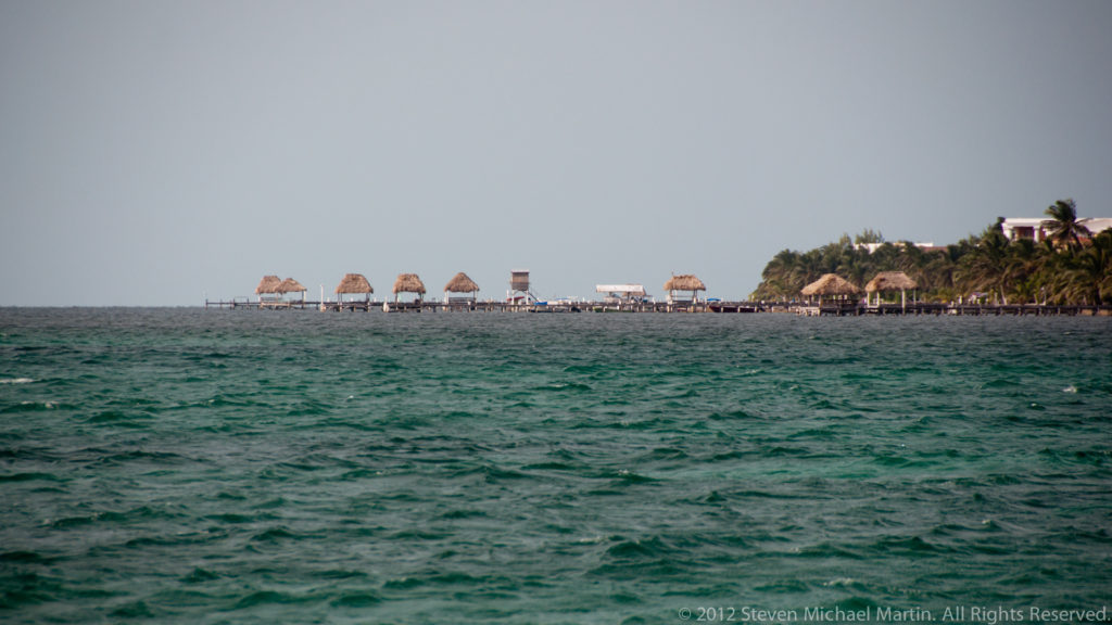 Choppy turquoise sea in the foreground with palm-lined Caye Caulker shoreline, low buildings, thatched shelters, and piers beyond.