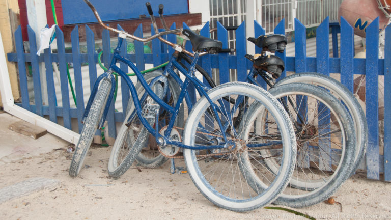Several blue bicycles with wide tires lean against a blue-and-white picket fence on a sandy street.