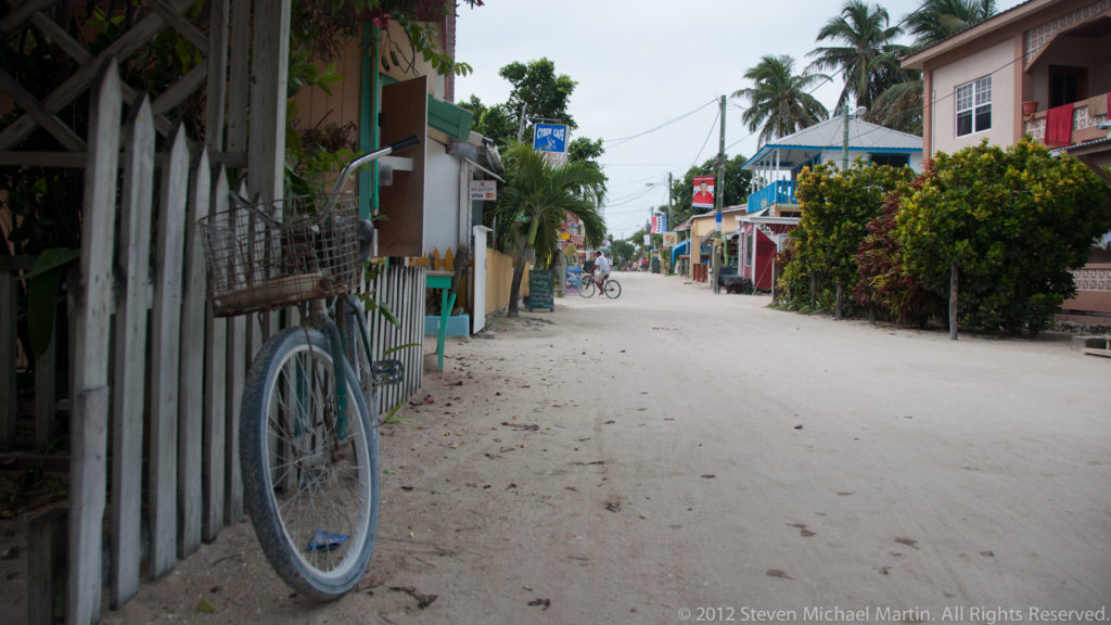 Sandy Caye Caulker street lined with houses, fences and tropical plants, with a bicycle in the foreground and cyclists in the distance.