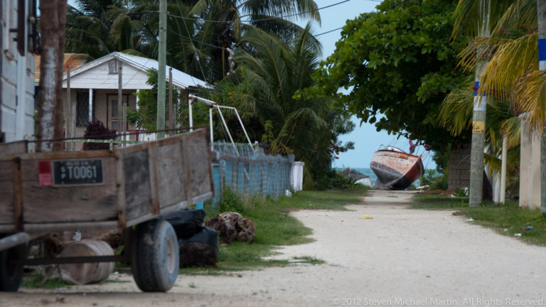 Sandy lane between homes and palms leading toward the sea and a boat, with a utility trailer in the foreground.