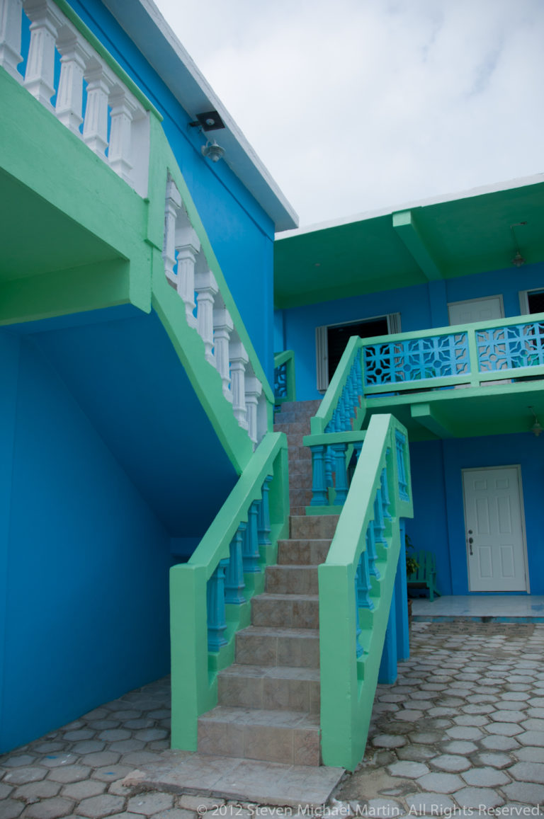 Turquoise courtyard building with mint-green twin stairways, decorative balcony rails, white doors, and a paved stone floor.