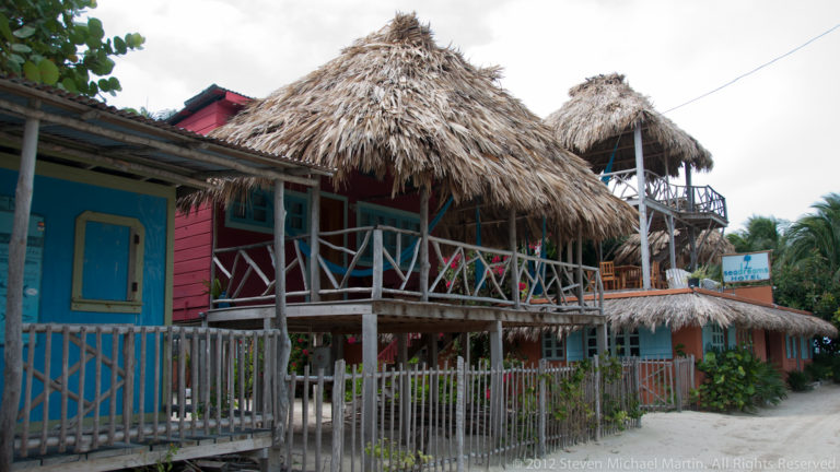Colorful raised wooden buildings with open verandas and steep thatched roofs beside a sandy lane on Caye Caulker.
