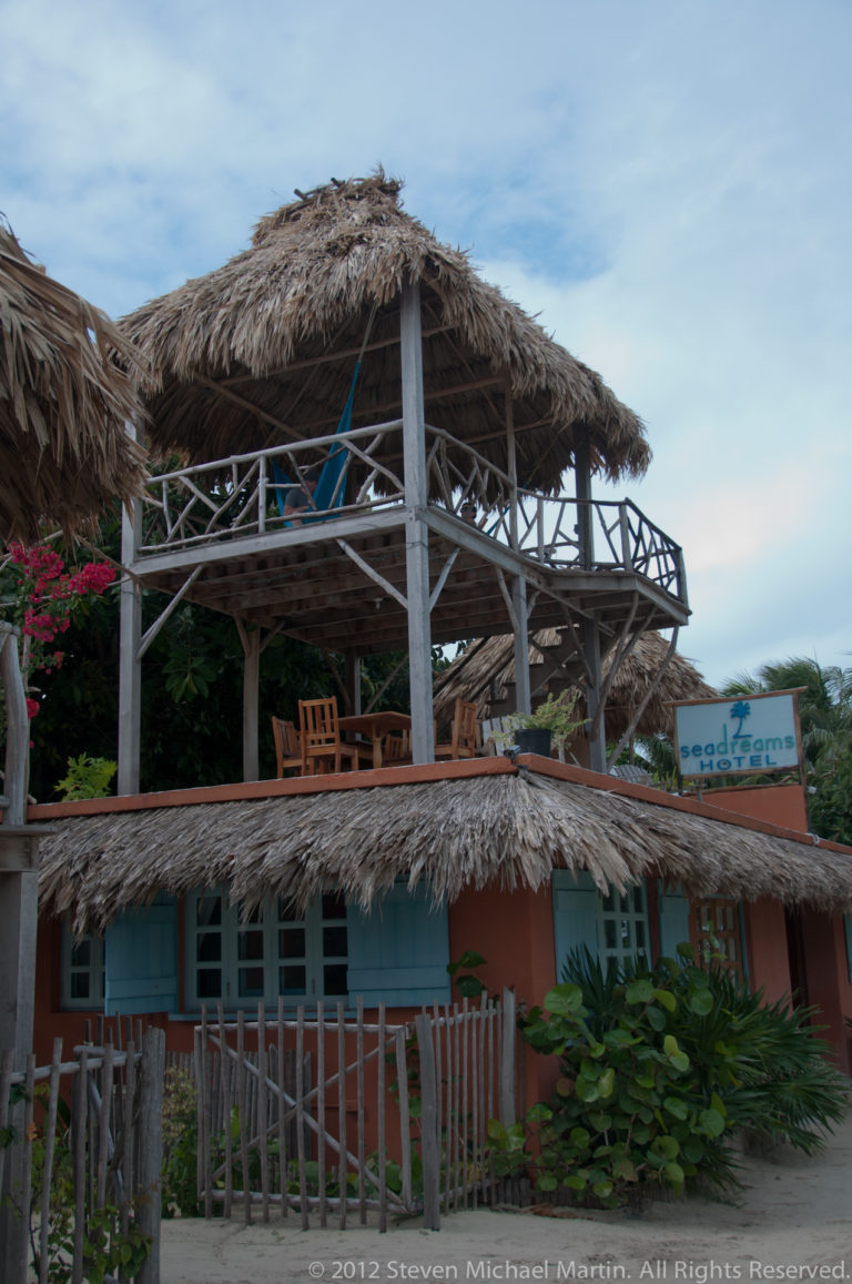 Two-level wooden building with broad thatched roofs, open upper deck, and cross-braced railings under a blue sky.